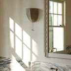 Unlit Clérachic linen wall sconce on a sunlit wall beside a framed window and a marble vanity with brass taps.