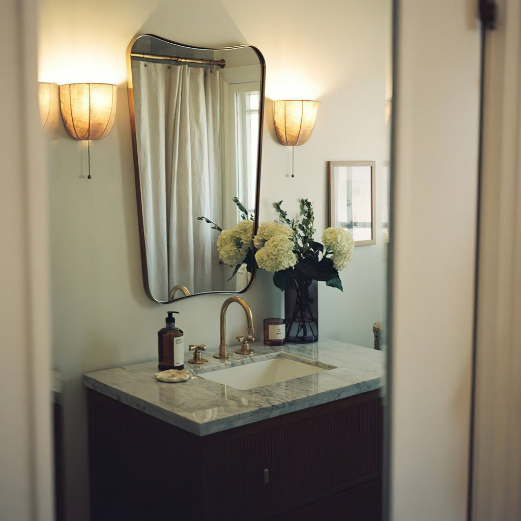 Two Clérachic linen wall sconces lit either side of a bathroom mirror above a marble-topped washstand with white hydrangeas.