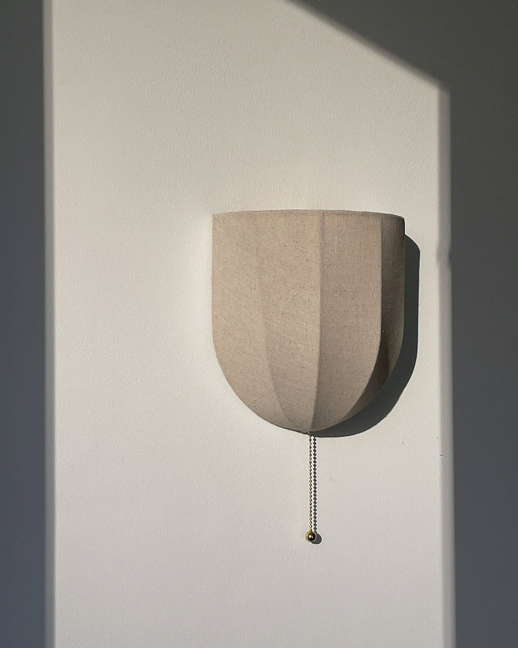 Unlit Clérachic linen wall sconce with a pull chain, on a white wall cut by a hard shadow.