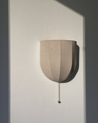 Unlit Clérachic linen wall sconce with a pull chain, on a white wall cut by a hard shadow.