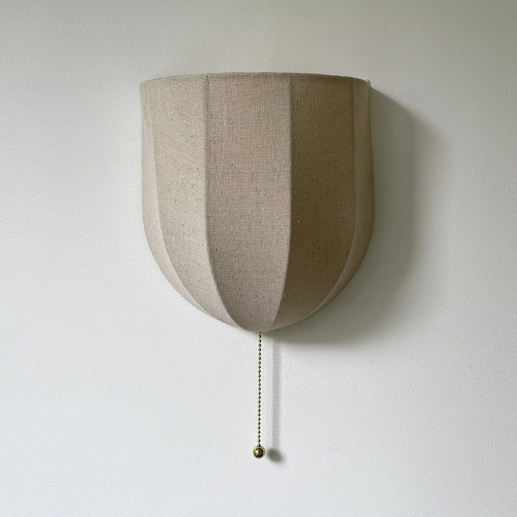 Close view of the vertical seams on this unlit Clérachic linen wall sconce, with a brass-toned pull chain on a pale wall.
