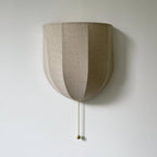 Close view of the vertical seams on this unlit Clérachic linen wall sconce, with a brass-toned pull chain on a pale wall.