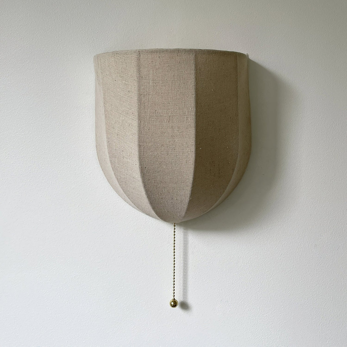 Close view of the vertical seams on this unlit Clérachic linen wall sconce, with a brass-toned pull chain on a pale wall.
