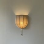 Lit Clérachic linen wall sconce glowing amber, light spilling upward over a grey wall and the shade's seams showing through.