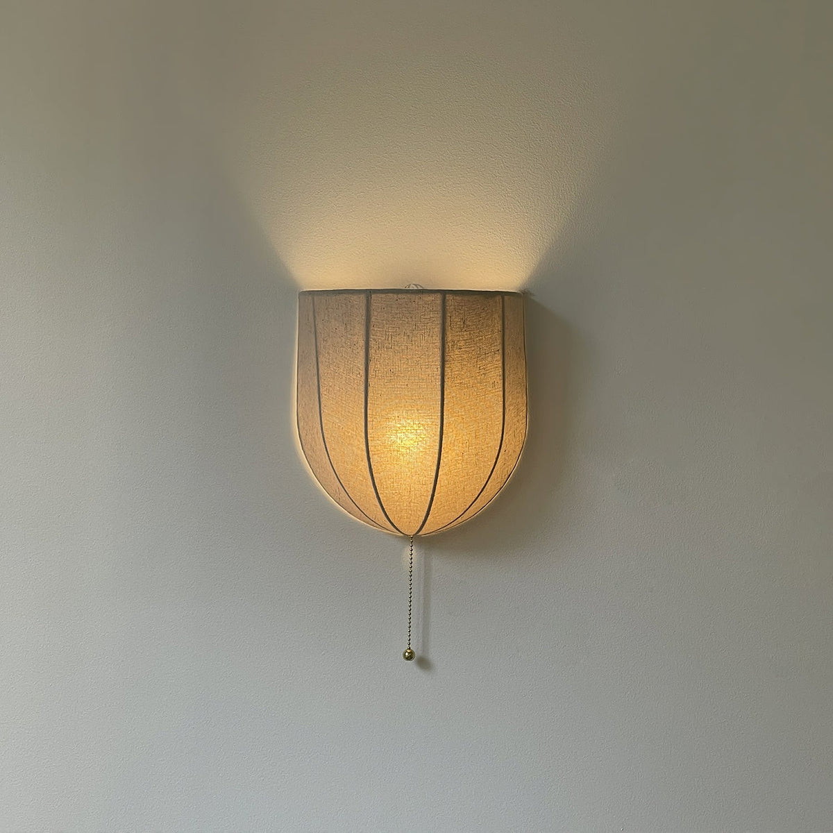Lit Clérachic linen wall sconce glowing amber, light spilling upward over a grey wall and the shade's seams showing through.