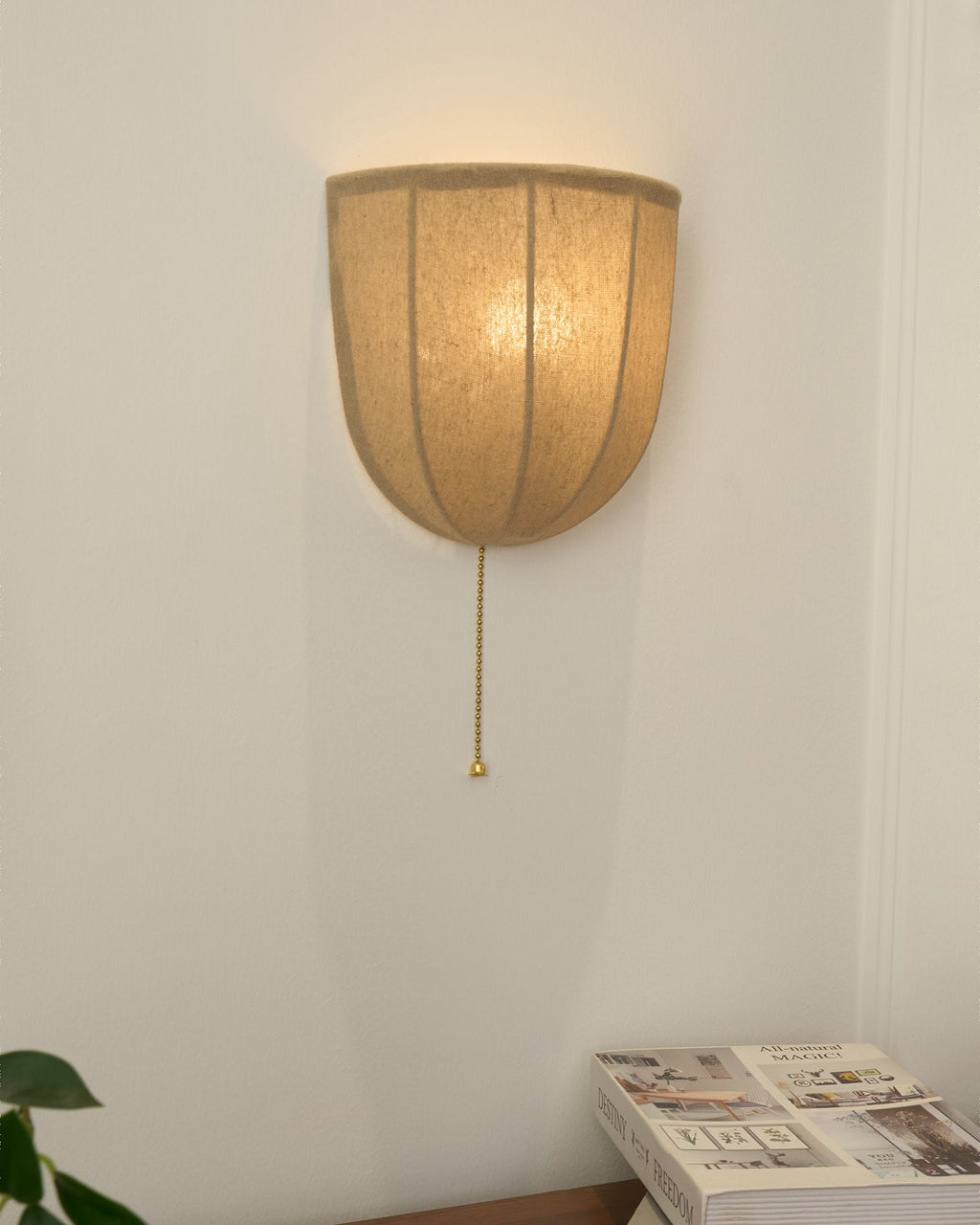 Lit Clérachic linen wall sconce on a white wall above a wooden sideboard, with magazines and green leaves nearby.