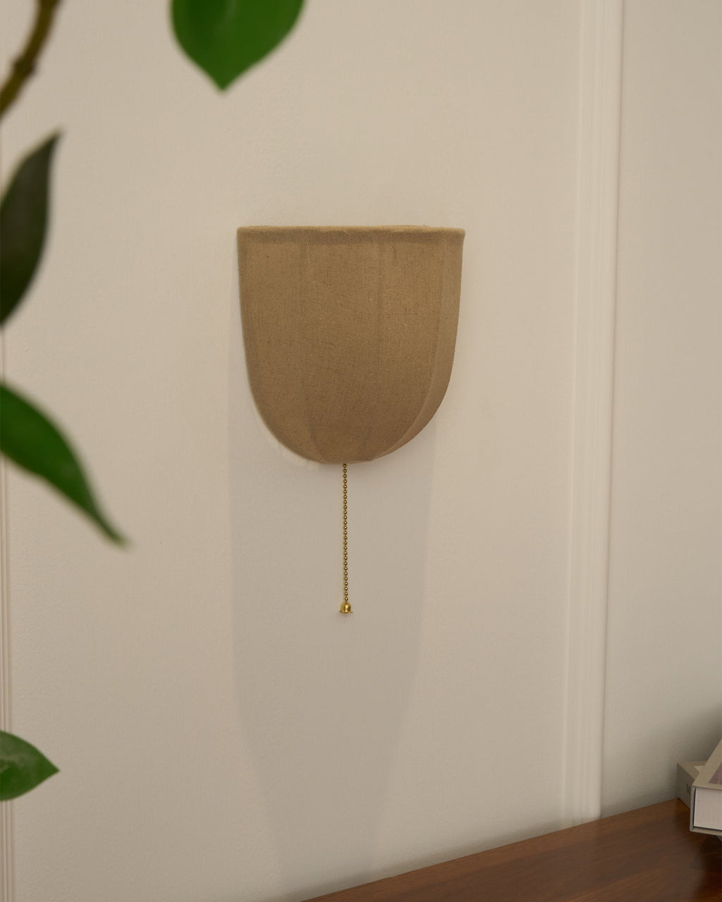 Unlit Clérachic linen wall sconce on a white wall, with large green leaves at the edge and a wooden surface below.