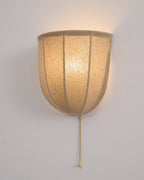 Close view of a lit Clérachic linen wall sconce, the seams dark against the glowing shade and a gold pull chain below.