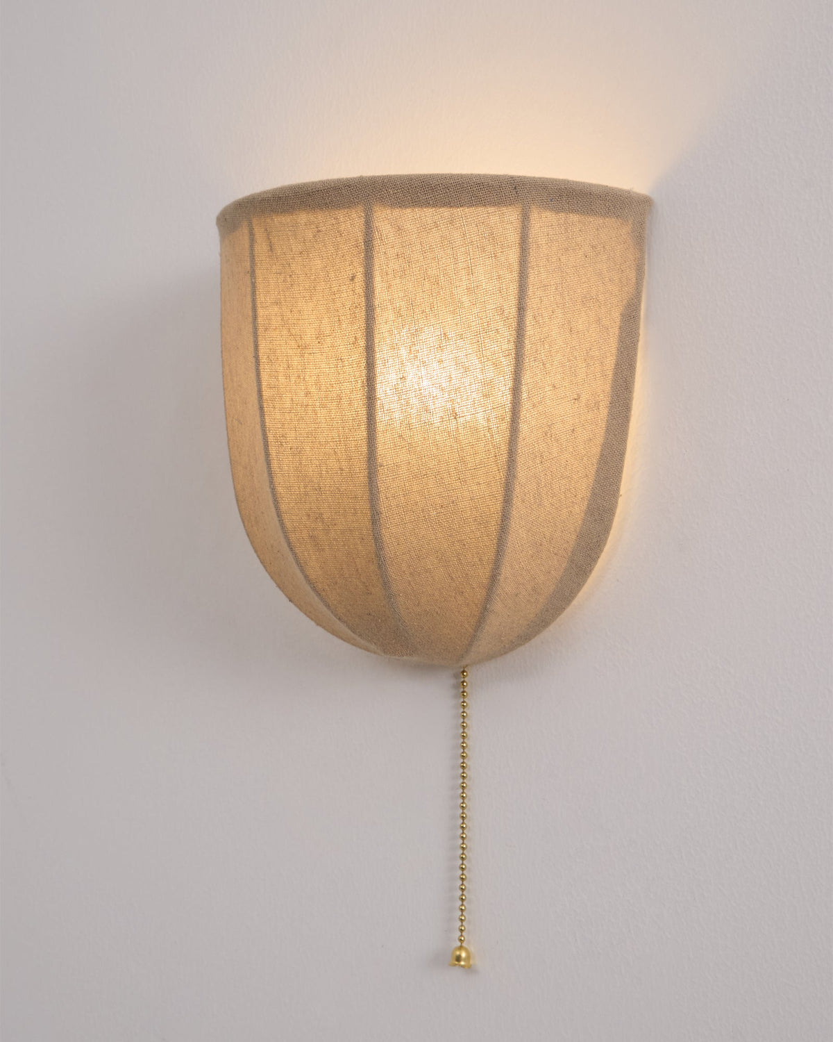 Close view of a lit Clérachic linen wall sconce, the seams dark against the glowing shade and a gold pull chain below.