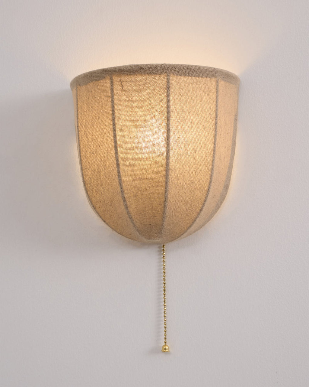 Lit Clérachic linen wall sconce on a pale wall, its ribbed shade glowing warm over a rim of natural linen.