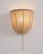 Lit Clérachic linen wall sconce on a pale wall, its ribbed shade glowing warm over a rim of natural linen.