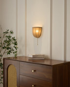 Clérachic linen wall sconce lit above a walnut chest with a cane door, a stack of books and a leafy branch.