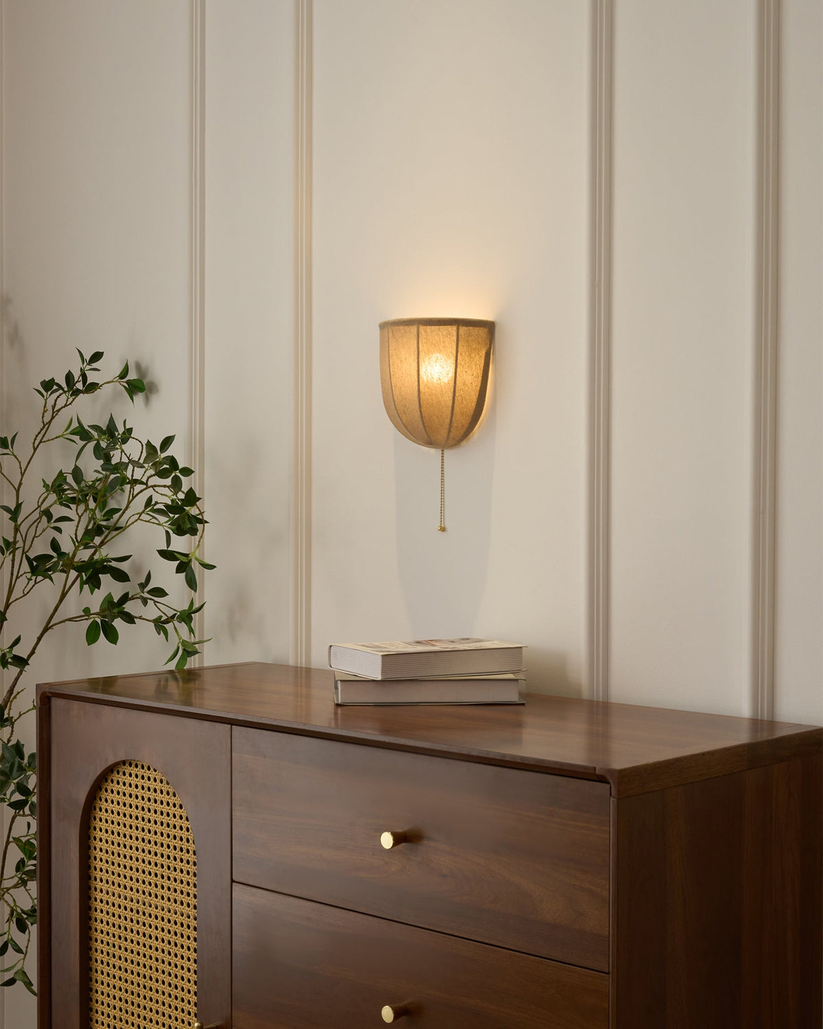Clérachic linen wall sconce lit above a walnut chest with a cane door, a stack of books and a leafy branch.