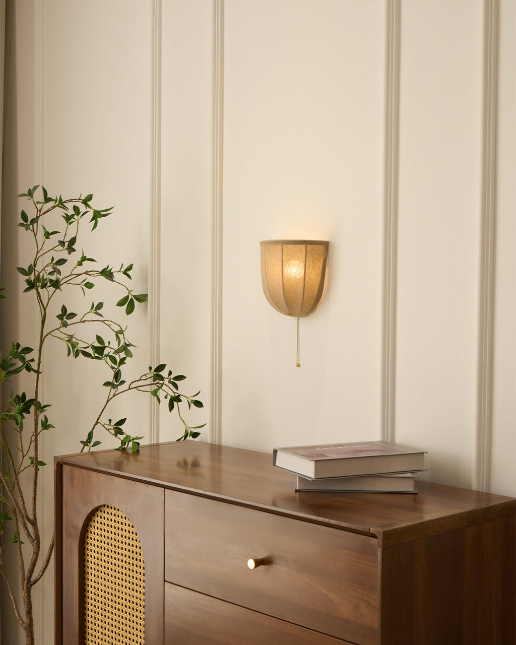 Lit Clérachic linen wall sconce on a ribbed panel wall, above a walnut cabinet with a brass handle and books.