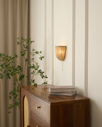 Lit Clérachic linen wall sconce on a panelled wall beside linen curtains, above a walnut cabinet and a leafy branch.