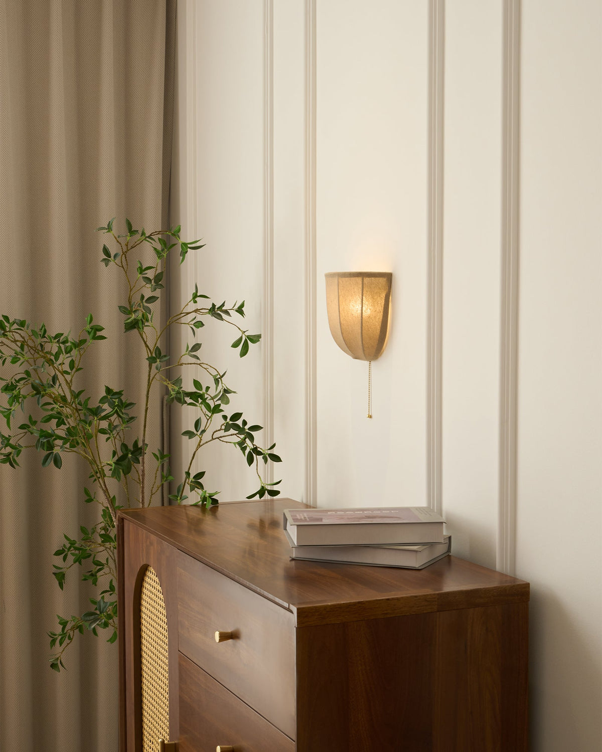 Lit Clérachic linen wall sconce on a panelled wall beside linen curtains, above a walnut cabinet and a leafy branch.