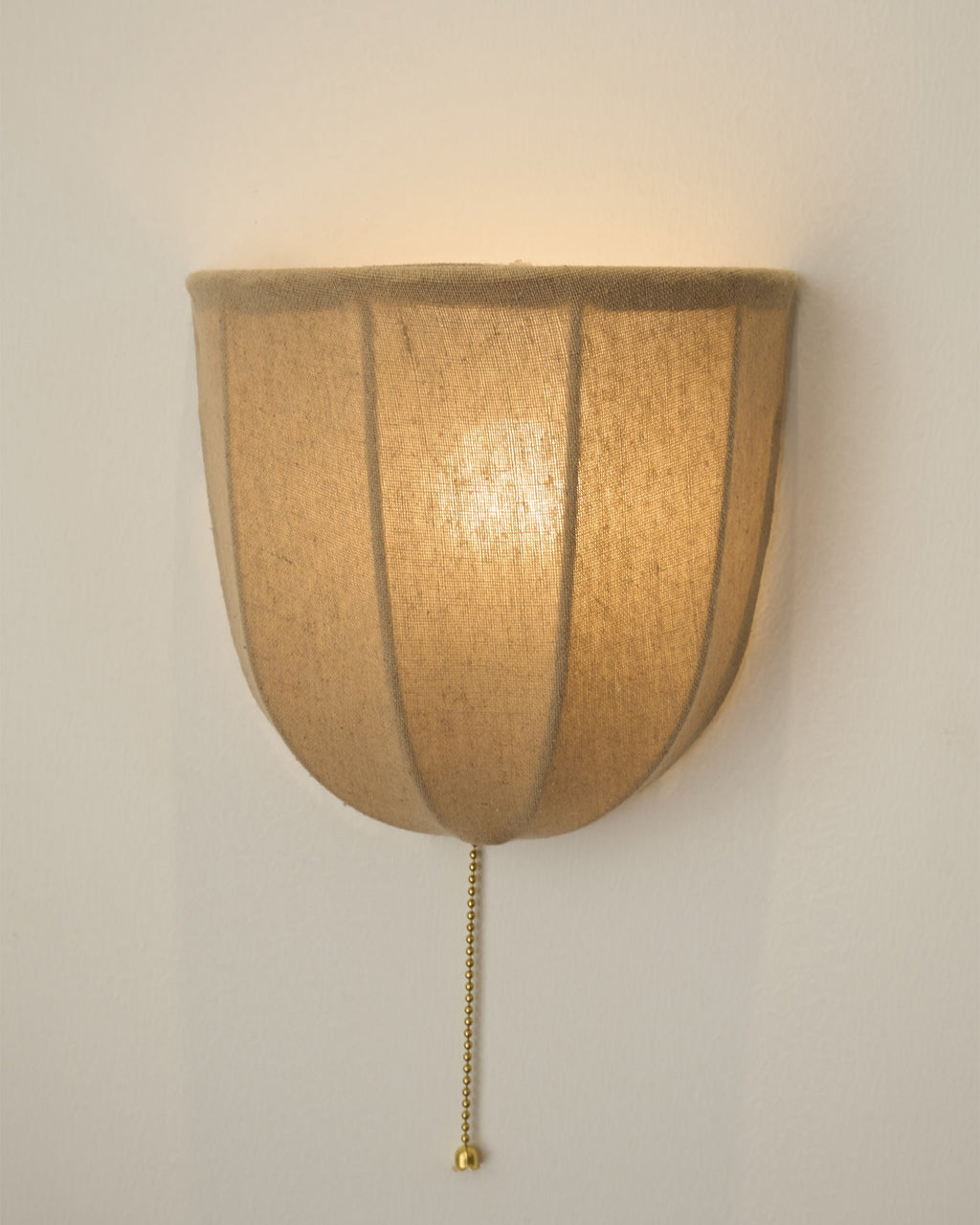 Large close view of a lit Clérachic linen wall sconce, with the glowing shade and a brass pull chain on a cream wall.