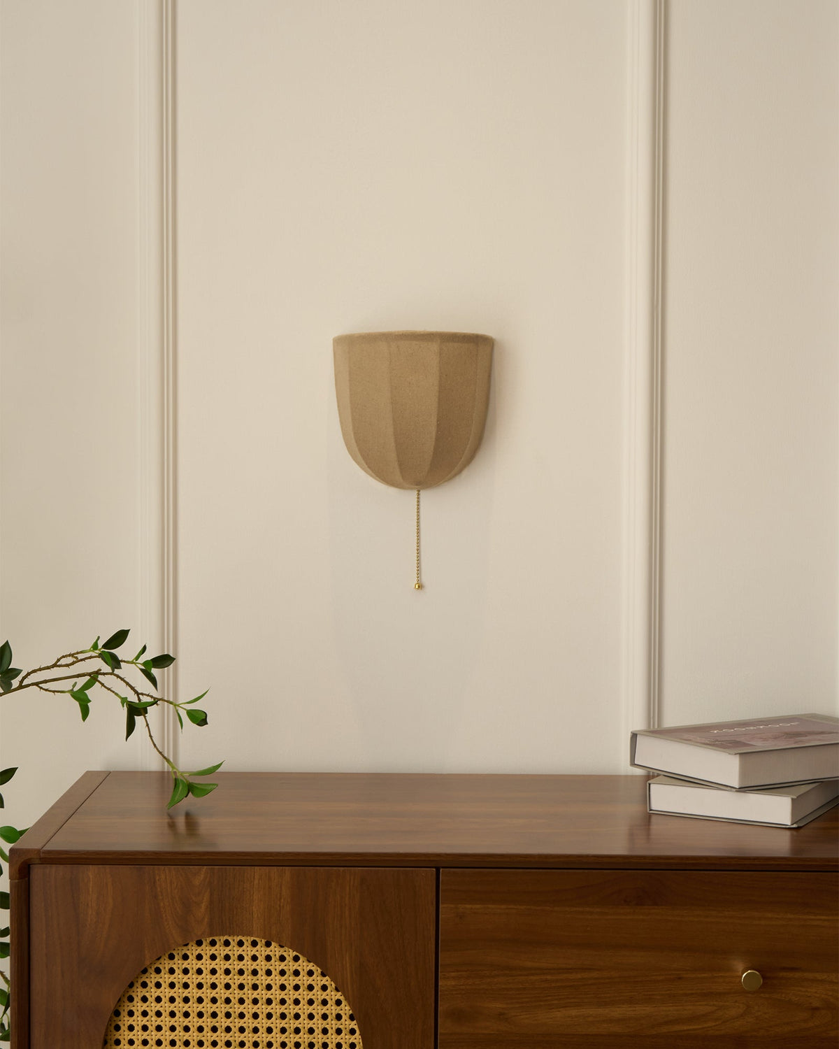 Small Clérachic linen wall sconce on a panelled cream wall above a walnut cabinet with a cane door and stacked books.