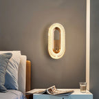 Clérachic gold oval wall lamp lit beside a bed, above a white bedside table with an open magazine and a glass.