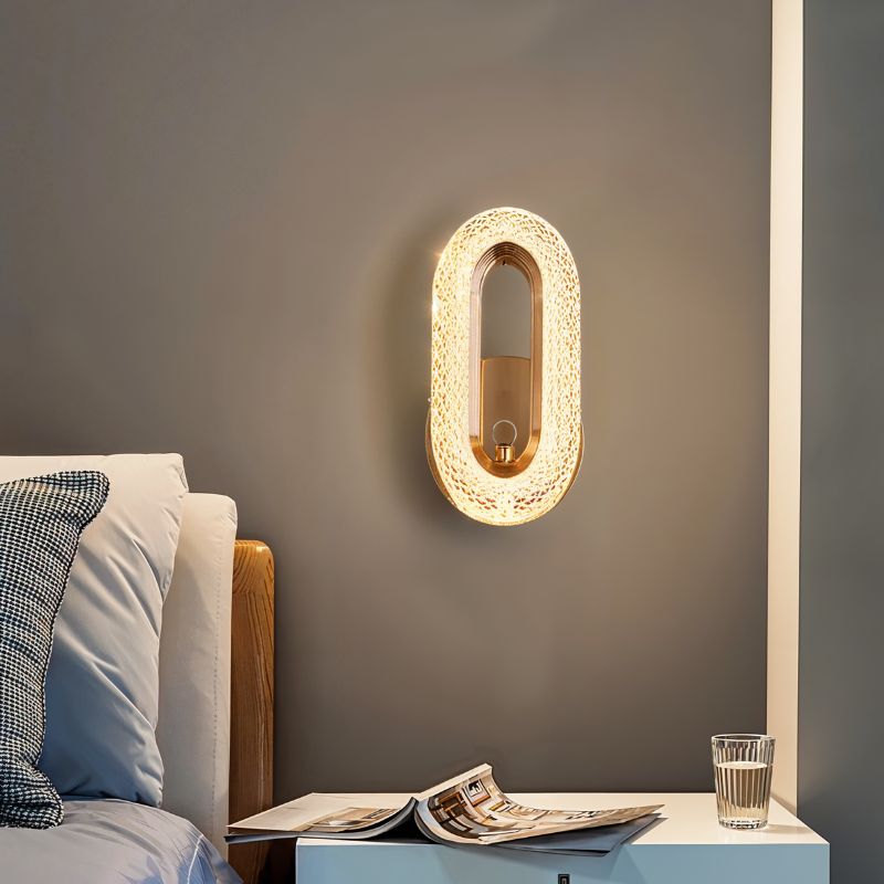 Clérachic gold oval wall lamp lit beside a bed, above a white bedside table with an open magazine and a glass.