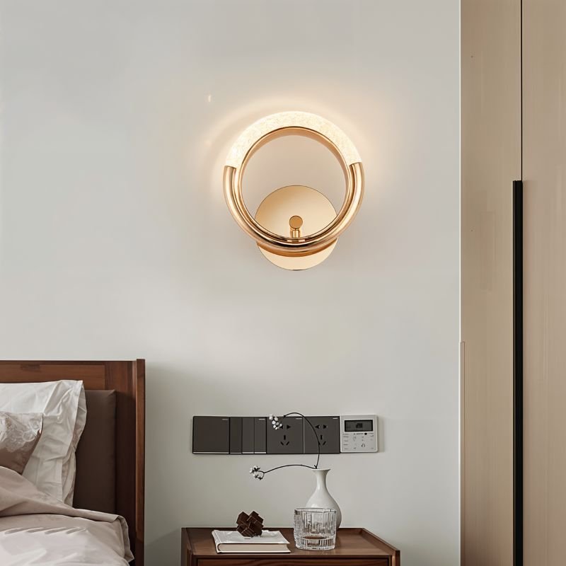 Clérachic gold ring wall lamp casting a soft glow on a pale wall above a wooden bedside table and a small vase.