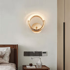 Clérachic gold ring wall lamp casting a soft glow on a pale wall above a wooden bedside table and a small vase.