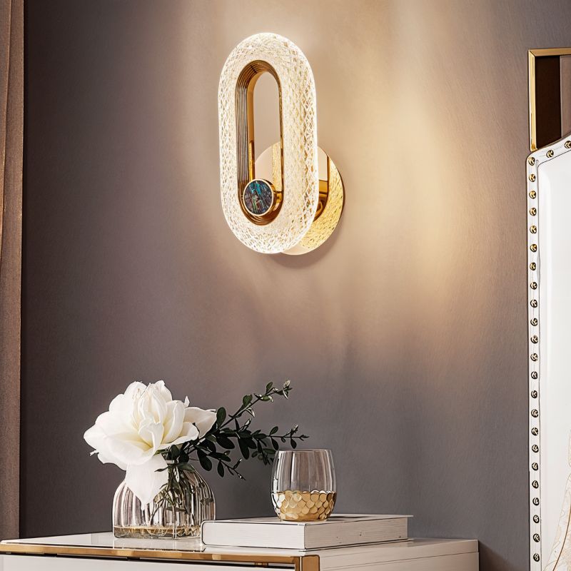 Clérachic gold oval wall lamp with a stone inlay lit on a taupe wall above a console with white flowers.