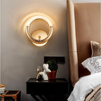 Clérachic gold ring wall lamp lit above a black bedside table with ornaments, next to a tan upholstered headboard.