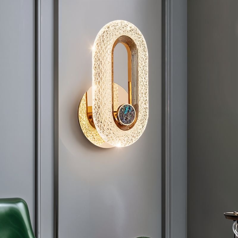 Clérachic gold oval wall lamp with a stone inlay, lit on a grey panelled wall beside a green leather chair.