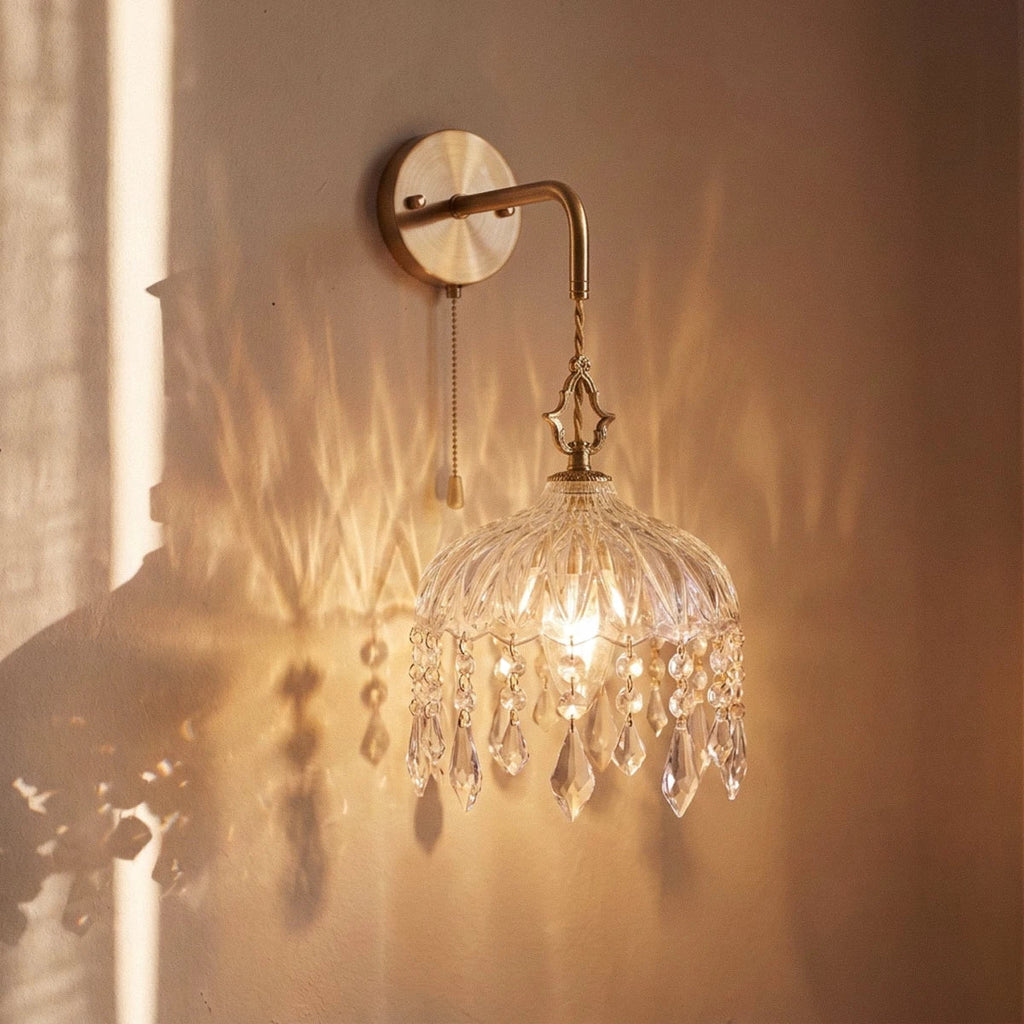 Clérachic gold wall lamp lit warm, its glass dome and crystal drops throwing patterned light across the wall.
