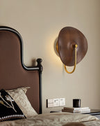 Beside a dark upholstered headboard, this Clérachic wall light shows a soft halo of warm light behind its walnut-colour wood disc.