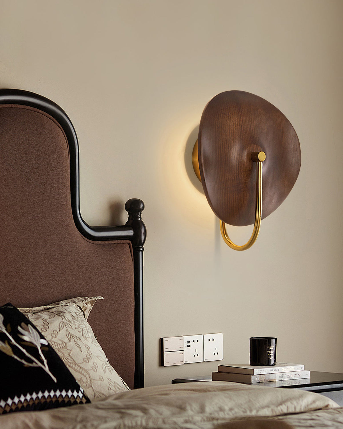 Beside a dark upholstered headboard, this Clérachic wall light shows a soft halo of warm light behind its walnut-colour wood disc.