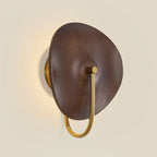 Front view of this Clérachic wall light, its walnut-colour disc lit warm from behind and its gold arm curving down on a cream wall.