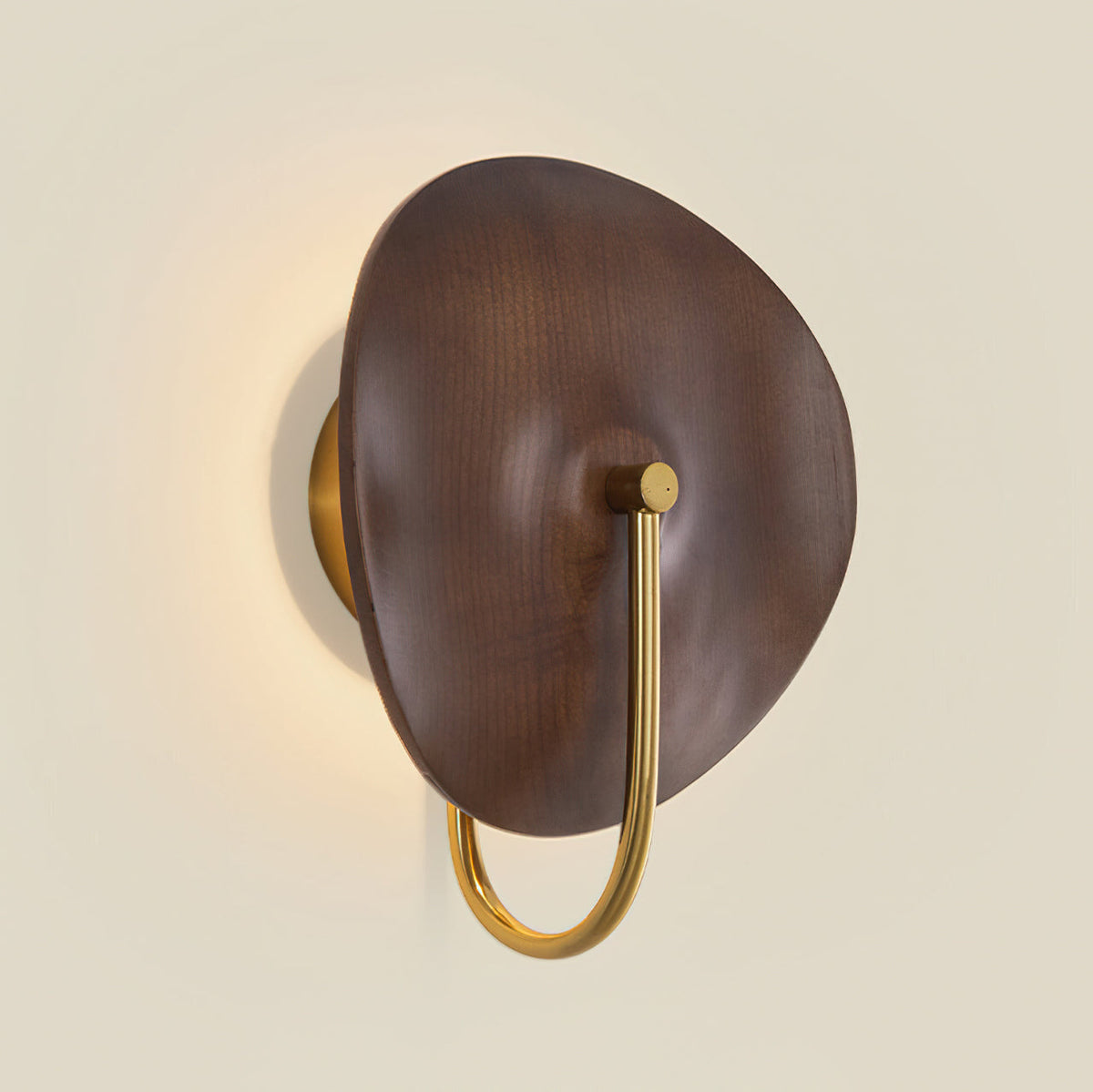 Front view of this Clérachic wall light, its walnut-colour disc lit warm from behind and its gold arm curving down on a cream wall.