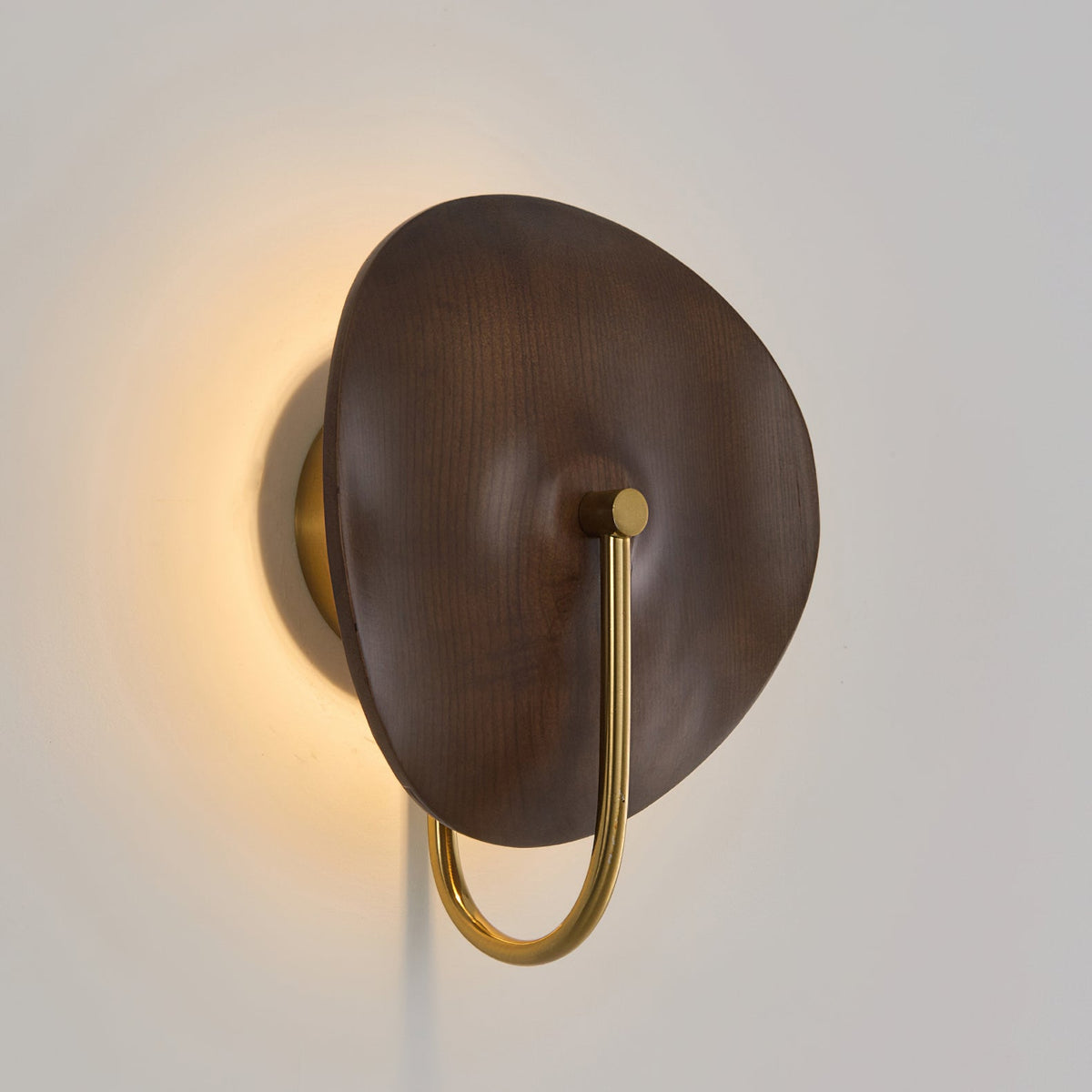 A soft amber glow spills around the edge of the walnut-colour wood disc on this Clérachic wall light, set against a pale grey wall.