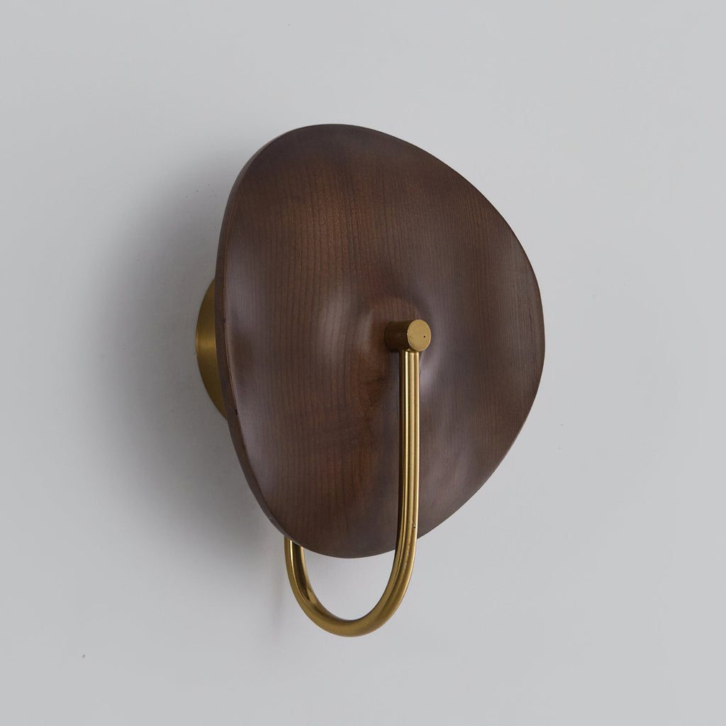 Three-quarter view of this unlit Clérachic wall light, with a rounded walnut-colour disc and a gold arm on a light grey wall.