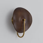 Three-quarter view of this unlit Clérachic wall light, with a rounded walnut-colour disc and a gold arm on a light grey wall.