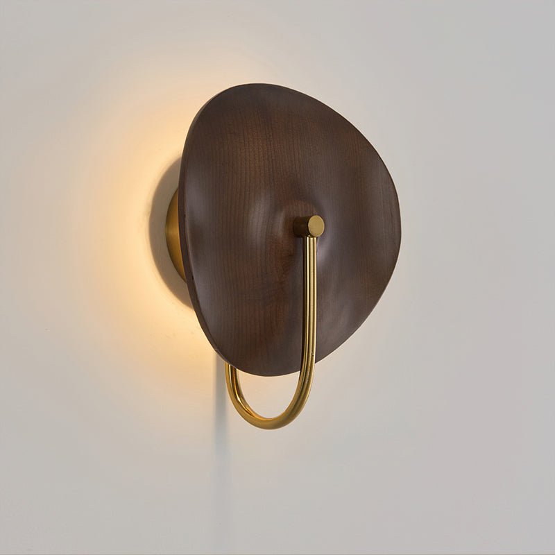 Angled view of this Clérachic wall light, a warm halo behind its dark walnut-colour disc and a gold arm below, on a grey wall.