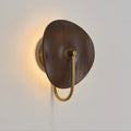 Angled view of this Clérachic wall light, a warm halo behind its dark walnut-colour disc and a gold arm below, on a grey wall.