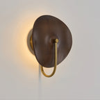 Angled view of this Clérachic wall light, a warm halo behind its dark walnut-colour disc and a gold arm below, on a grey wall.