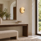 A small round Clérachic wall sconce glows on a cream wall beside an arched mirror, above a dark wood console and a boucle bench.
