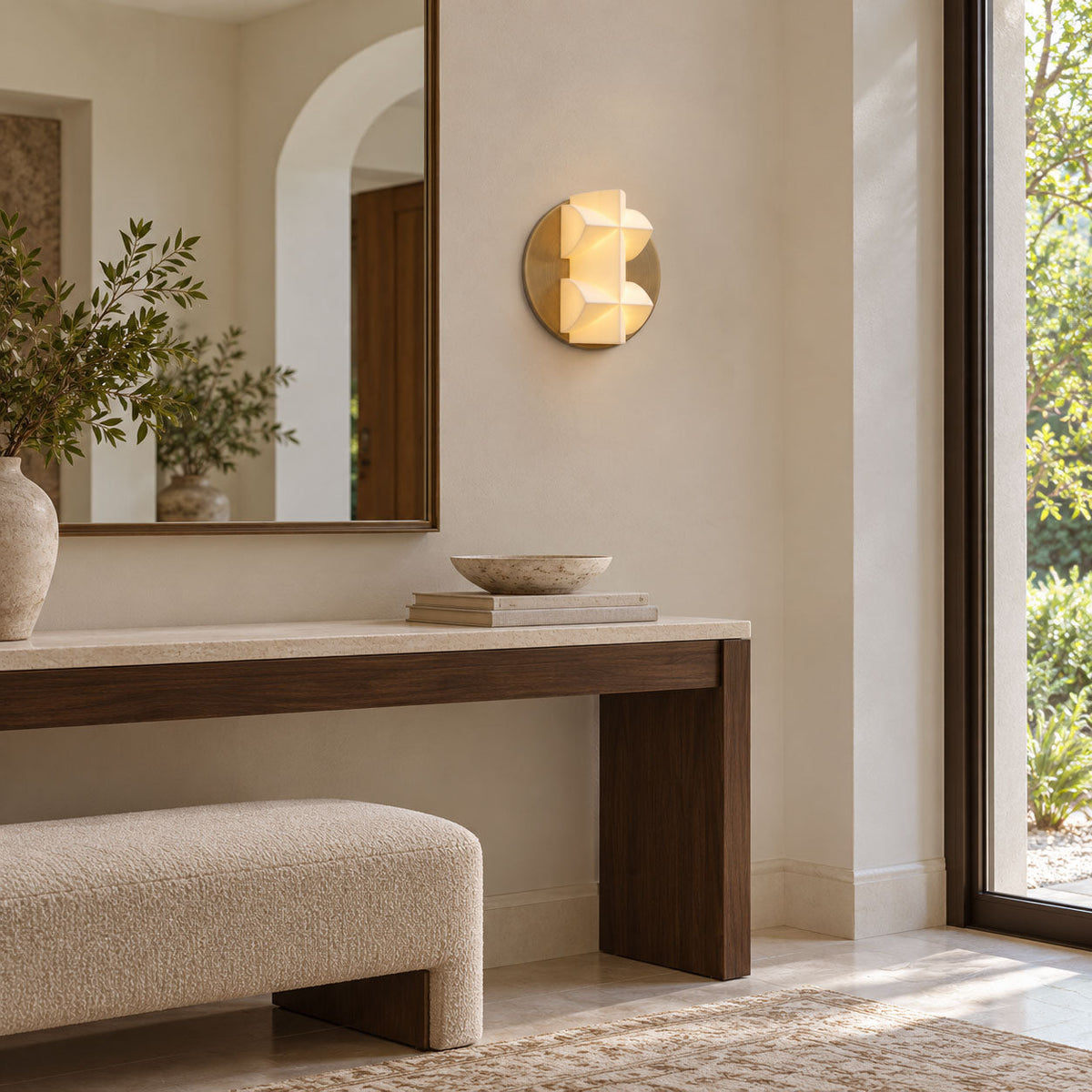 A small round Clérachic wall sconce glows on a cream wall beside an arched mirror, above a dark wood console and a boucle bench.