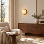 Lit gold-and-white Clérachic sconce on a warm neutral wall, with an armchair, a walnut sideboard and a vase of olive branches.