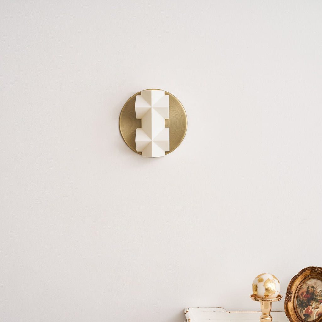 Unlit, this Clérachic wall sconce shows its white faceted shade on a brushed gold disc, against a pale wall above a small white table.