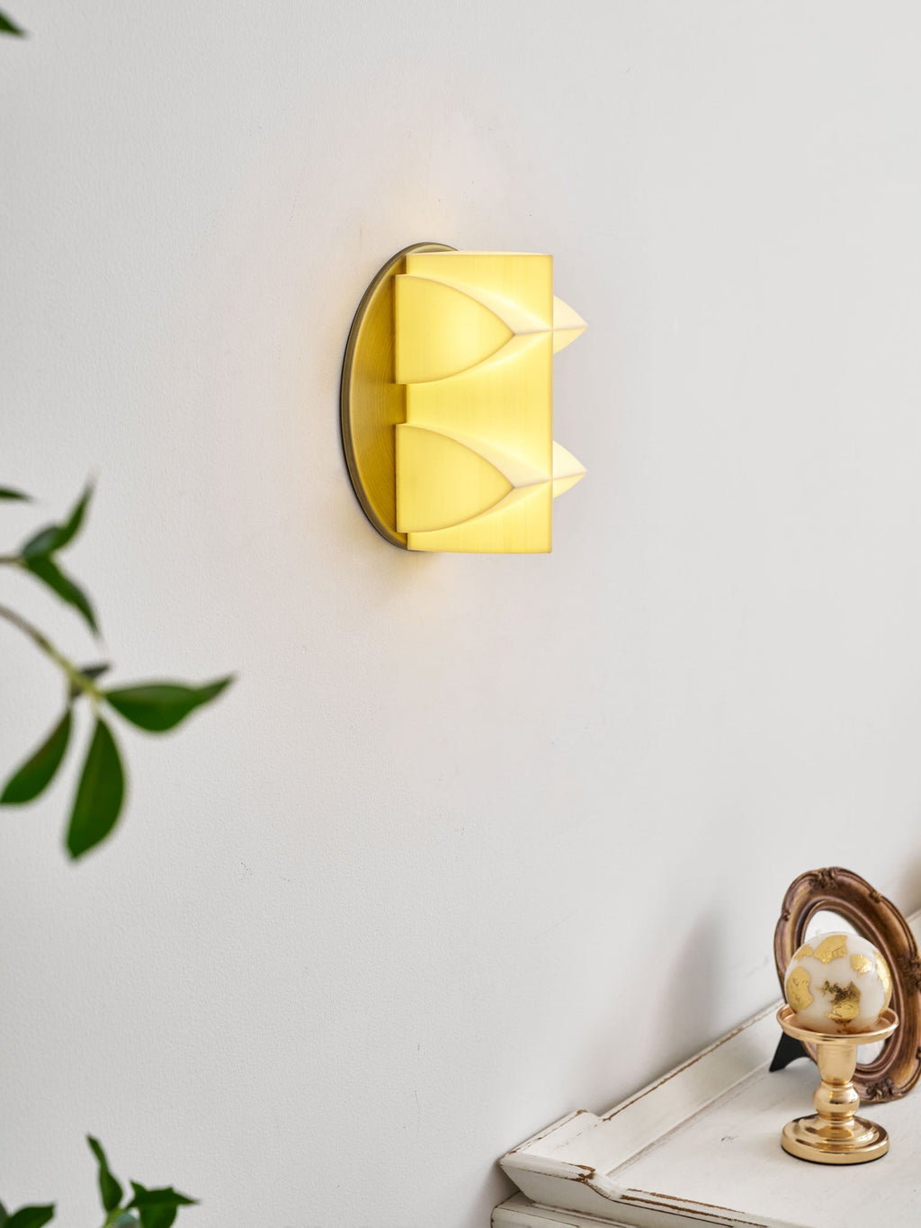 Angled view of a Clérachic sconce lit a soft yellow, its curved points catching the glow, with a gold ornament on a table below.