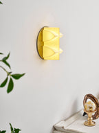 Angled view of a Clérachic sconce lit a soft yellow, its curved points catching the glow, with a gold ornament on a table below.