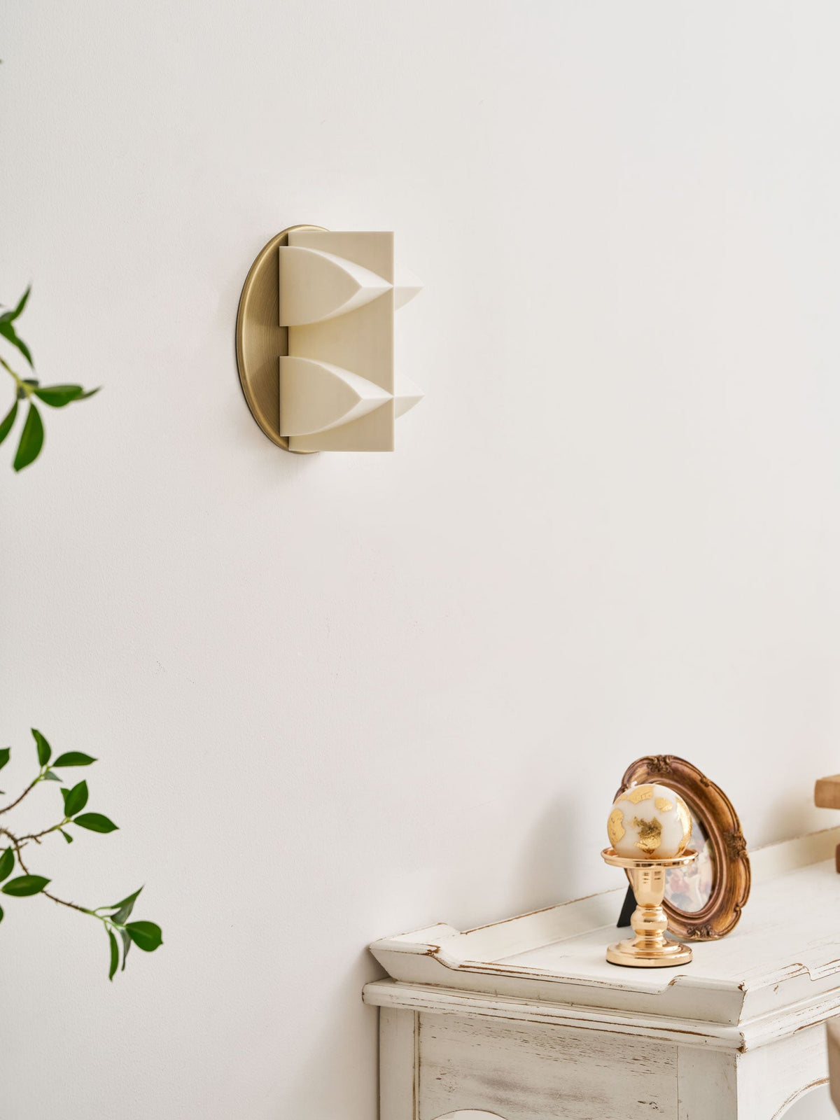 Side view of an unlit cream Clérachic wall sconce, its two curved points projecting from a gold disc, with a leafy branch at the left.