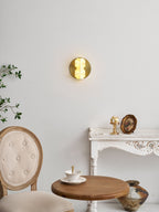 A small lit Clérachic sconce on a pale wall above a white carved table, a round wooden table and a tufted chair.