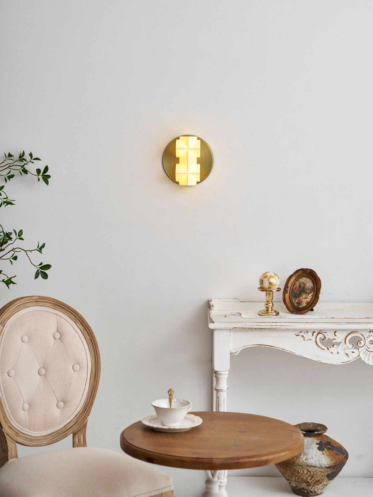 A small lit Clérachic sconce on a pale wall above a white carved table, a round wooden table and a tufted chair.