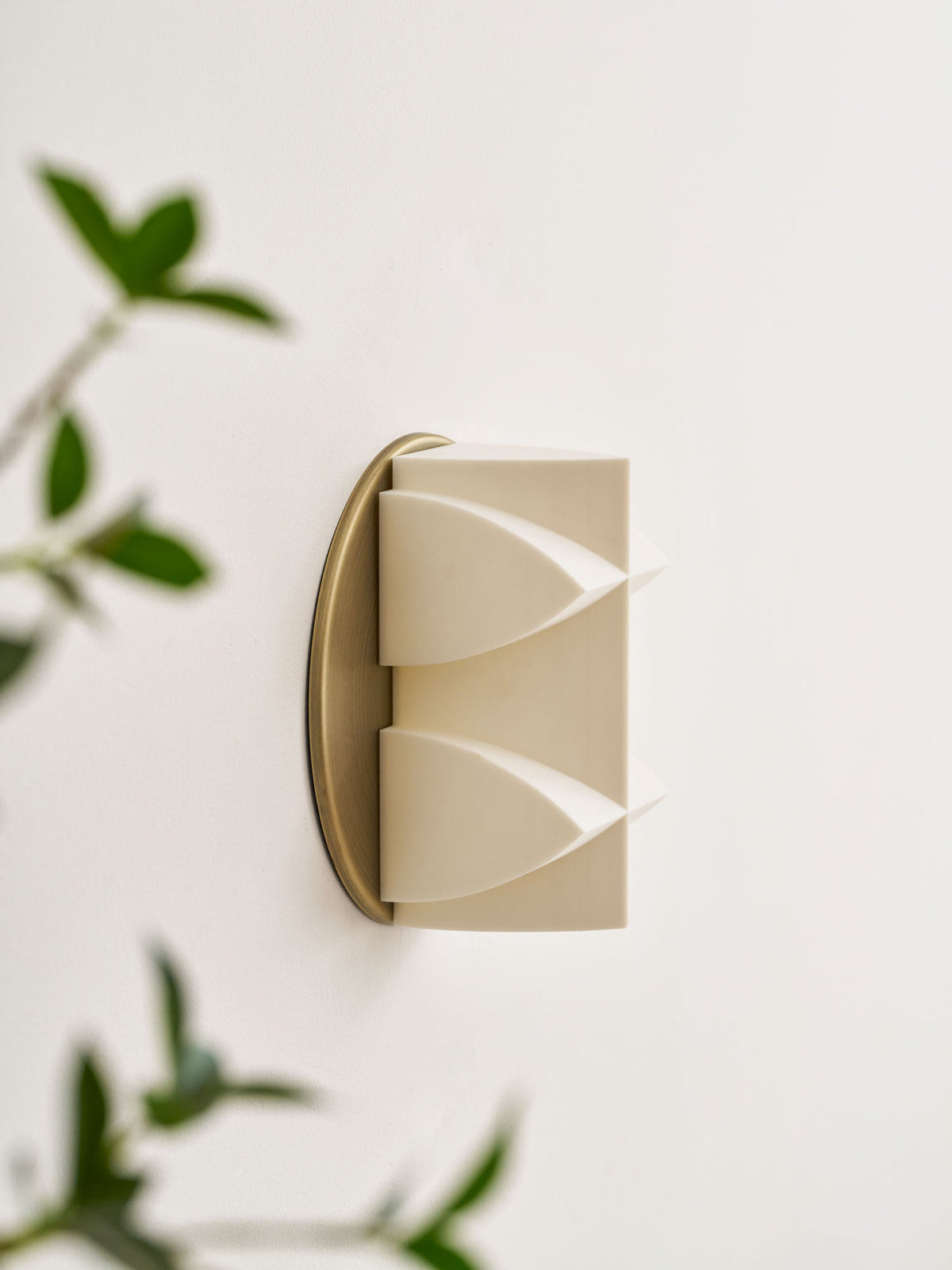Close, angled view of an unlit cream Clérachic sconce on a gold disc, with blurred green leaves on either side.